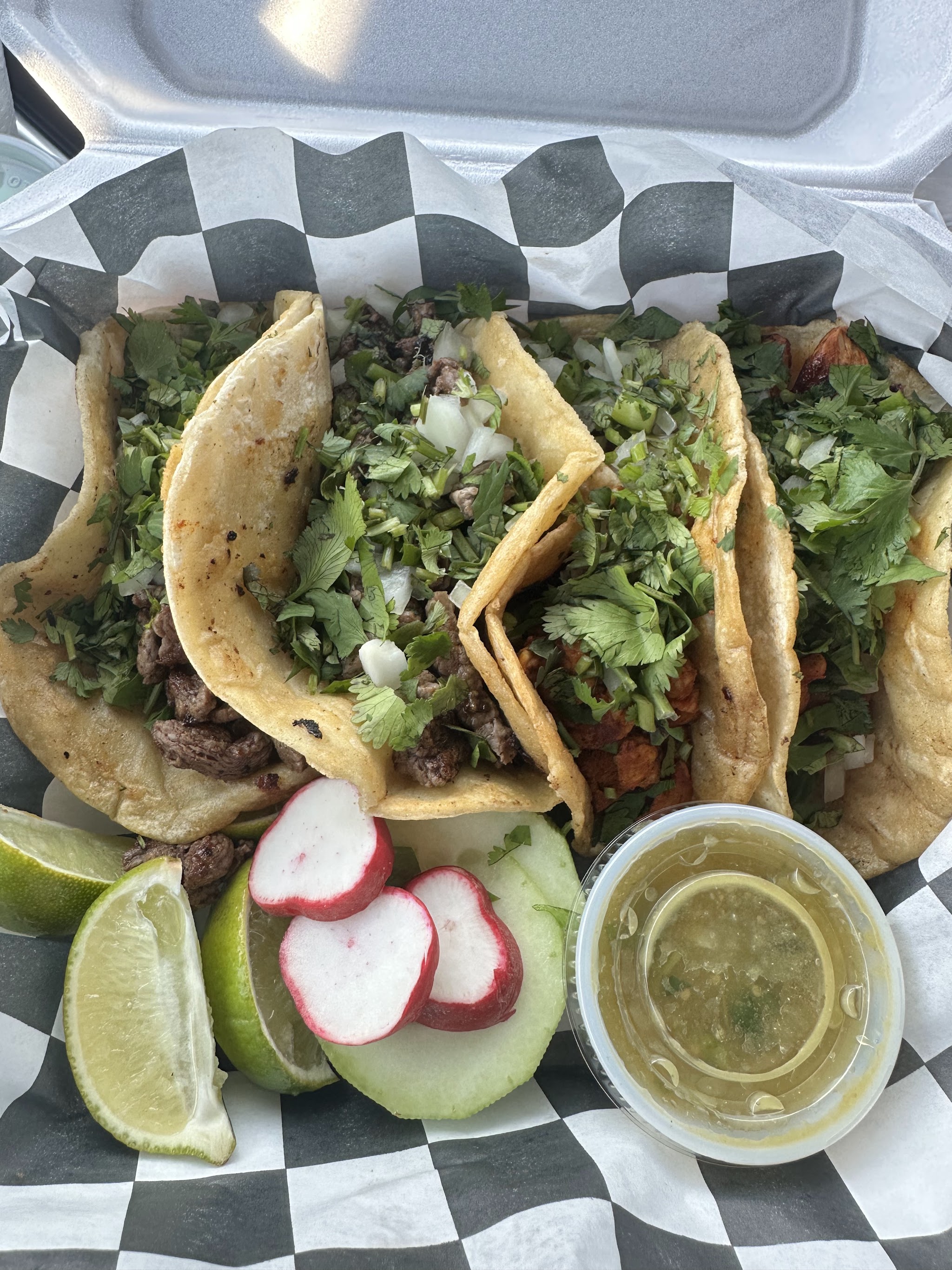 Five street tacos in checkered paper with lime wedges, radish and a cup of green salsa
