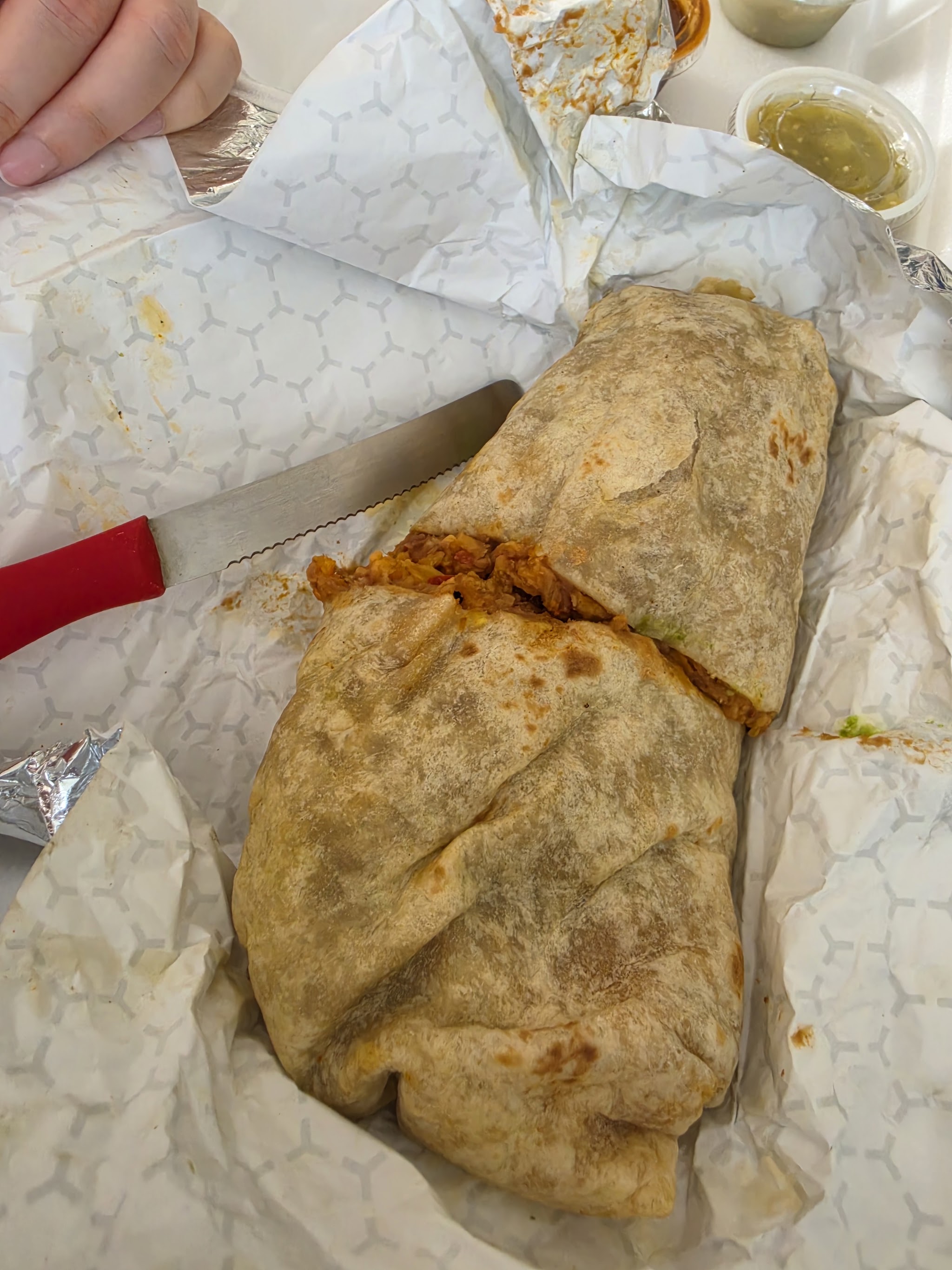 A big burrito cut in half next to cups of green salsa